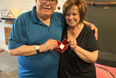 Smiling couple holding a red stained-glass heart ornament together in a cozy craft workshop with rustic decor