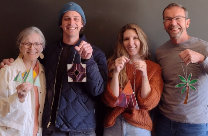 Four smiling people holding handmade stained-glass ornaments — green sailboat, purple geometric square, orange triangular teepee, and green palm tree — at a casual DIY stained-glass craft workshop against a dark wall.