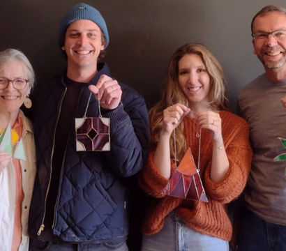 Four smiling people holding handmade stained-glass ornaments — green sailboat, purple geometric square, orange triangular teepee, and green palm tree — at a casual DIY stained-glass craft workshop against a dark wall.