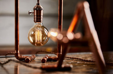 Warm-glowing Edison filament bulb dangling inside a copper-pipe industrial table lamp on a rustic wooden surface.