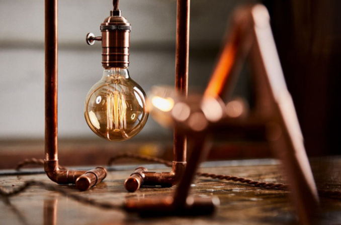 Warm-glowing Edison filament bulb dangling inside a copper-pipe industrial table lamp on a rustic wooden surface.
