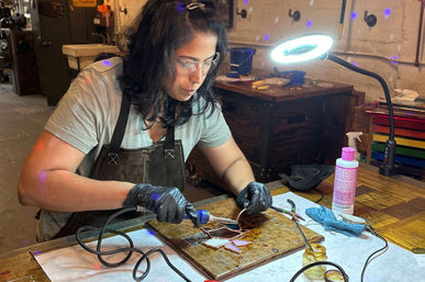 Artist soldering stained-glass pieces at a wooden workbench in a studio workshop, wearing safety glasses and gloves, with a gooseneck lamp, soldering iron, pliers and flux bottle nearby.