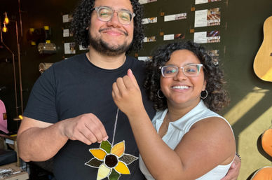 Two smiling people wearing glasses hold a handmade stained-glass sunflower ornament in a sunlit craft studio with guitars on the wall
