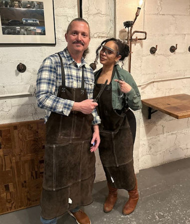 Two smiling people in a rustic industrial workshop wearing leather aprons and boots, one holding a handcrafted knife while the other points to it beside a wooden workbench and exposed brick wall.