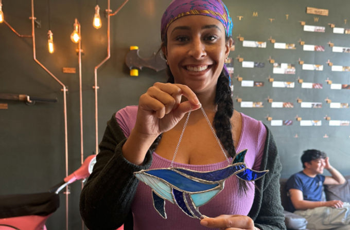 Person smiling and holding a blue stained-glass whale suncatcher in a cozy cafe with hanging Edison bulbs and a photo display wall
