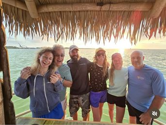 Six adults smiling and posing under a thatched-roof tiki hut on a wooden dock, overlooking calm green coastal water at sunset with a seagull flying in the background.