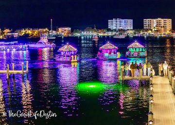 Nighttime coastal marina with three tiki-roof boats covered in colorful LED lights reflecting purple and green on the water, spectators gathered on a wooden pier and illuminated buildings along the shoreline.