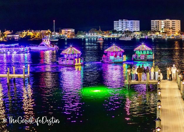 Nighttime coastal marina with three tiki-roof boats covered in colorful LED lights reflecting purple and green on the water, spectators gathered on a wooden pier and illuminated buildings along the shoreline.