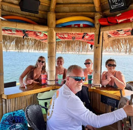 Group of smiling adults in swimsuits at a bamboo floating tiki bar on calm water, captain at the helm steering and colorful drink cups on the wooden bar.