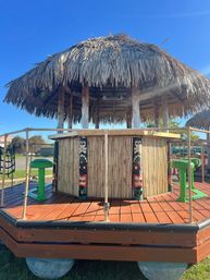 Tiki-style thatched-roof outdoor bar on a raised wooden deck, decorated with colorful tiki totem posts and bright green stools under a clear blue sky.