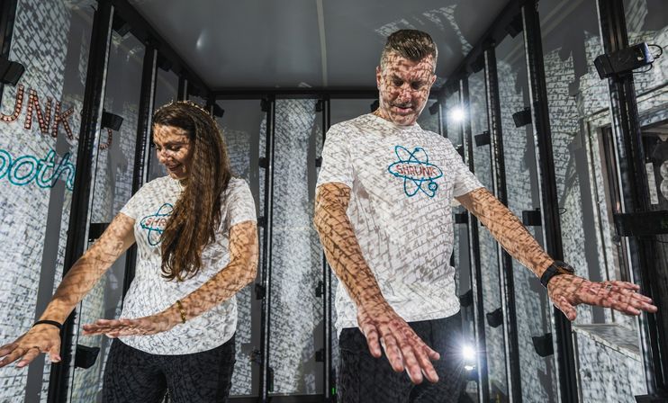 Two visitors in an indoor immersive projection light installation, patterned text and grid-like shadows projected across their white shirts and glass panels as they reach forward inside the exhibit.