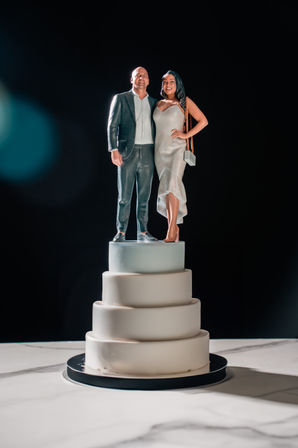 Chic four-tier white wedding cake topped with a realistic couple figurine — man in a suit and woman in a silver dress with a small purse, displayed on a marble table against a black backdrop.