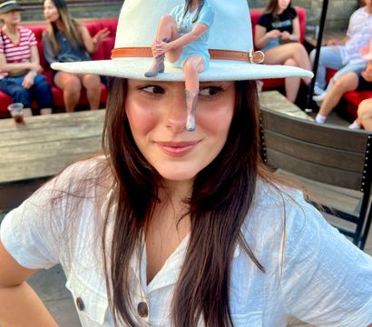 Smiling woman on an urban rooftop patio wearing a white cowboy hat topped by a tiny seated figurine on the brim