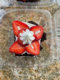 Close-up top view of a chocolate cupcake topped with a swirl of whipped cream and fresh sliced strawberries in a clear plastic takeout container on a speckled granite countertop