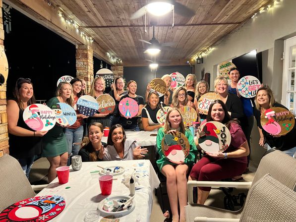 Smiling group of women and girls on a covered backyard patio holding round painted holiday signs at a festive DIY craft night under string lights.