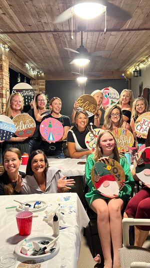 Large group of smiling women at a festive holiday craft night on a covered patio, each holding decorated round wooden signs under string lights and ceiling fans