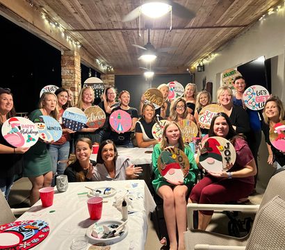 Large group of smiling women at a festive holiday craft night on a covered patio, each holding decorated round wooden signs under string lights and ceiling fans