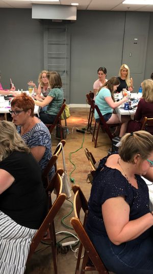 Busy indoor workshop room with a group of women seated at long tables doing DIY nail-art and crafts, surrounded by tools, polish bottles, cups and snacks.