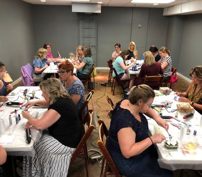 Hands-on DIY craft workshop in a community room — women seated at tables with paints, glue guns and tools, chatting and working on small projects