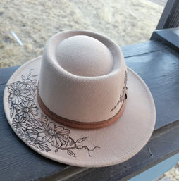 Beige felt western cowboy hat with a brown leather band and hand-drawn black floral design on the brim, resting on a dark wooden railing with dry grass in the background — rustic outdoor vibe.
