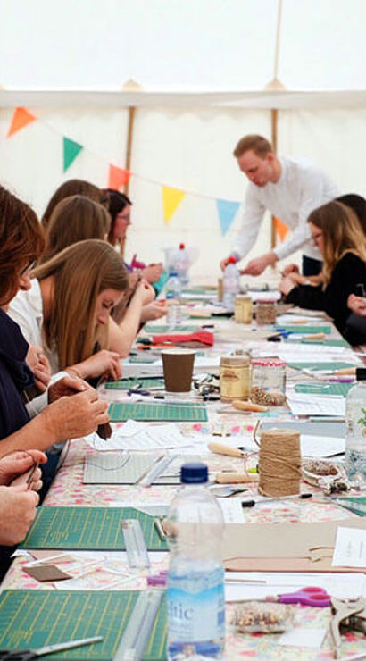 Community DIY crafting workshop inside a white marquee, people seated along a long table with cutting mats, paper, twine and tools, colorful bunting overhead