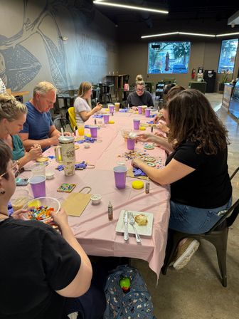Adults gathered around a long pink-covered table in a cozy cafe-style studio making colorful mosaic coasters with small tiles, glue bottles and purple cups nearby.