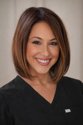 Smiling female healthcare professional headshot with a brown bob haircut, natural makeup, and black scrub top against a soft neutral background — friendly clinic staff portrait.