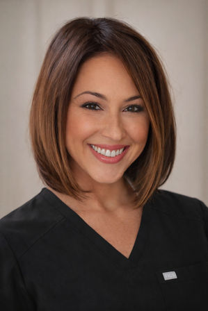 Smiling female healthcare professional headshot with a brown bob haircut, natural makeup, and black scrub top against a soft neutral background — friendly clinic staff portrait.