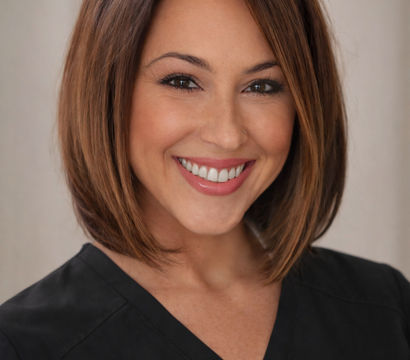 Smiling female healthcare professional headshot with a brown bob haircut, natural makeup, and black scrub top against a soft neutral background — friendly clinic staff portrait.