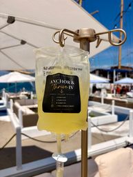Yellow IV hydration bag hanging on a stand at a sunny seaside lounge with white umbrellas, cabanas and sailboat masts in the background.