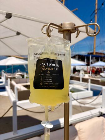 Yellow IV hydration bag hanging on a stand at a sunny seaside lounge with white umbrellas, cabanas and sailboat masts in the background.