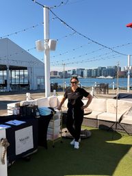 Smiling person in black uniform at a sunny waterfront event lounge with string lights, outdoor sofas, tables, and a city skyline and boats across the harbor