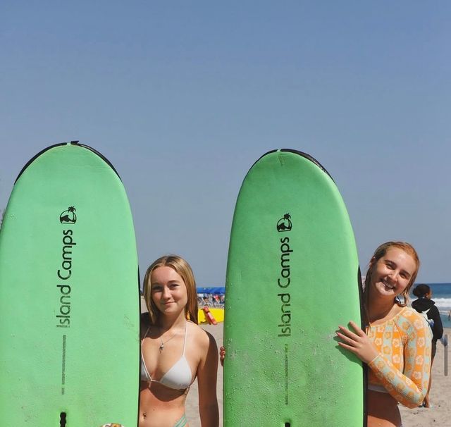 Two smiling young women holding bright green surfboards on a sunny ocean beach with clear blue sky and gentle waves in the background.