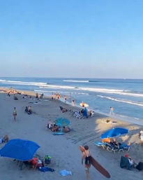 Busy summer sandy beach with surfers carrying boards, blue umbrellas and sunbathers along the shoreline with gentle ocean waves under a clear blue sky