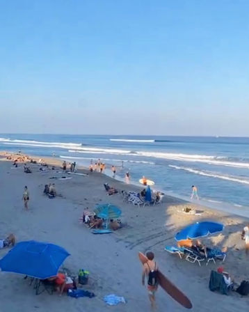 Busy summer sandy beach with surfers carrying boards, blue umbrellas and sunbathers along the shoreline with gentle ocean waves under a clear blue sky