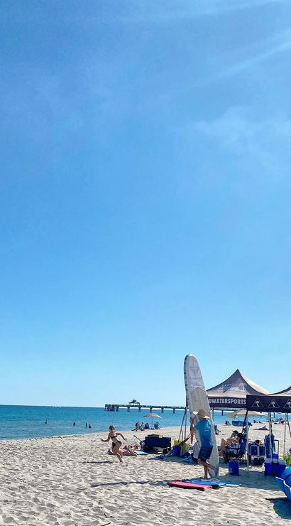 Sunny oceanfront beach under a clear blue sky, people playing near watersports tents and surfboards with a pier in the distance.