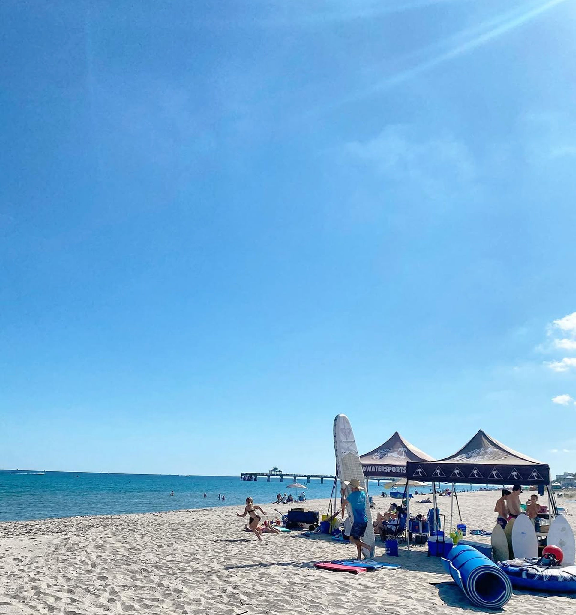 Sunny oceanfront beach under a clear blue sky, people playing near watersports tents and surfboards with a pier in the distance.