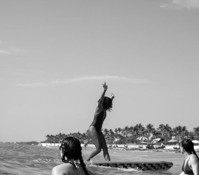 Surfer balancing on a longboard with arms raised in shallow ocean waves, two swimmers watching and a palm-lined tropical beach with umbrellas in the background.
