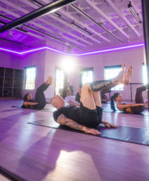 Adults in a purple-lit indoor fitness studio performing Pilates-style core leg lifts on mats during a group class