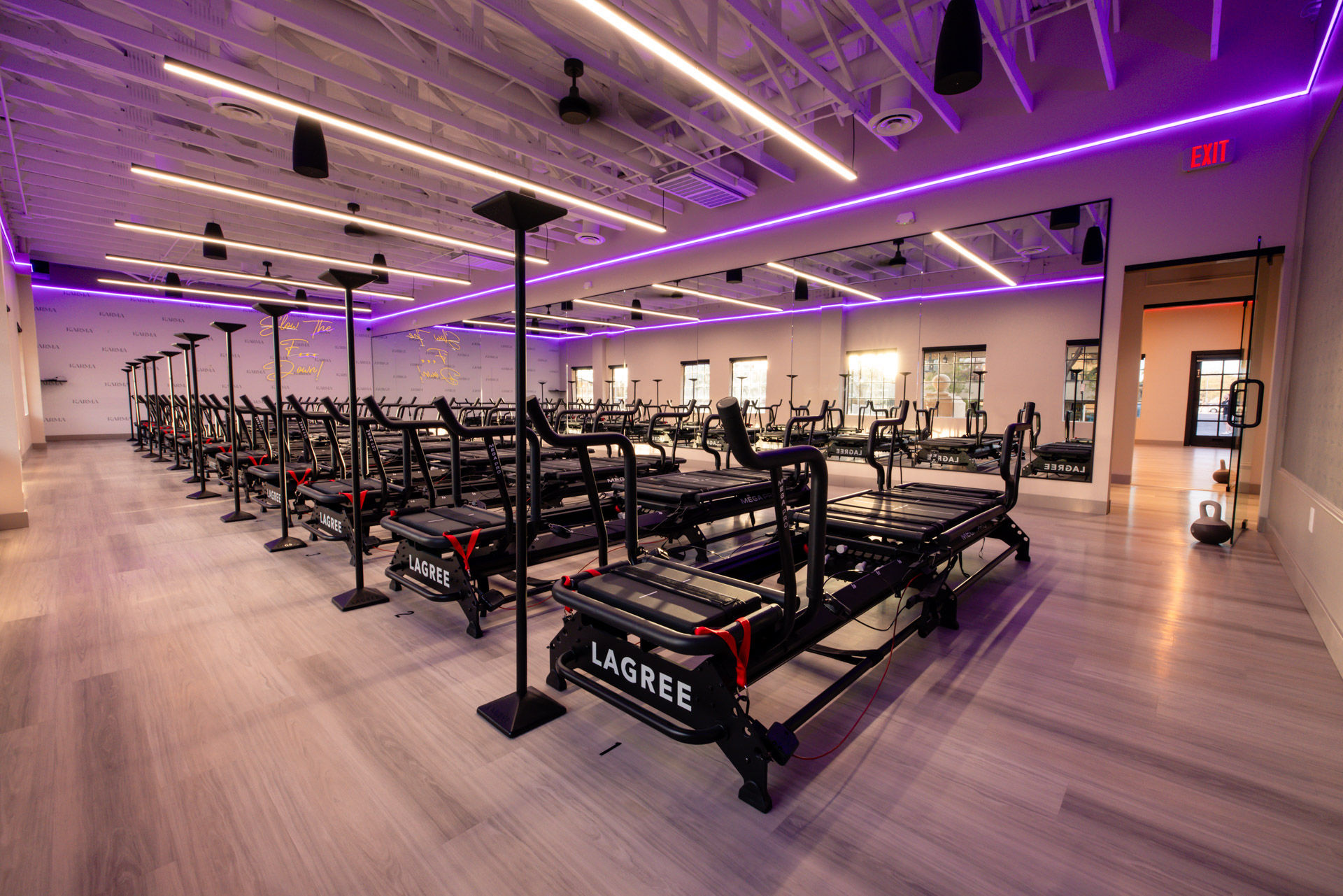 Modern, spacious pilates studio interior with rows of reformer machines, purple LED ceiling lights, mirrored wall and light wood floors ready for a group workout.