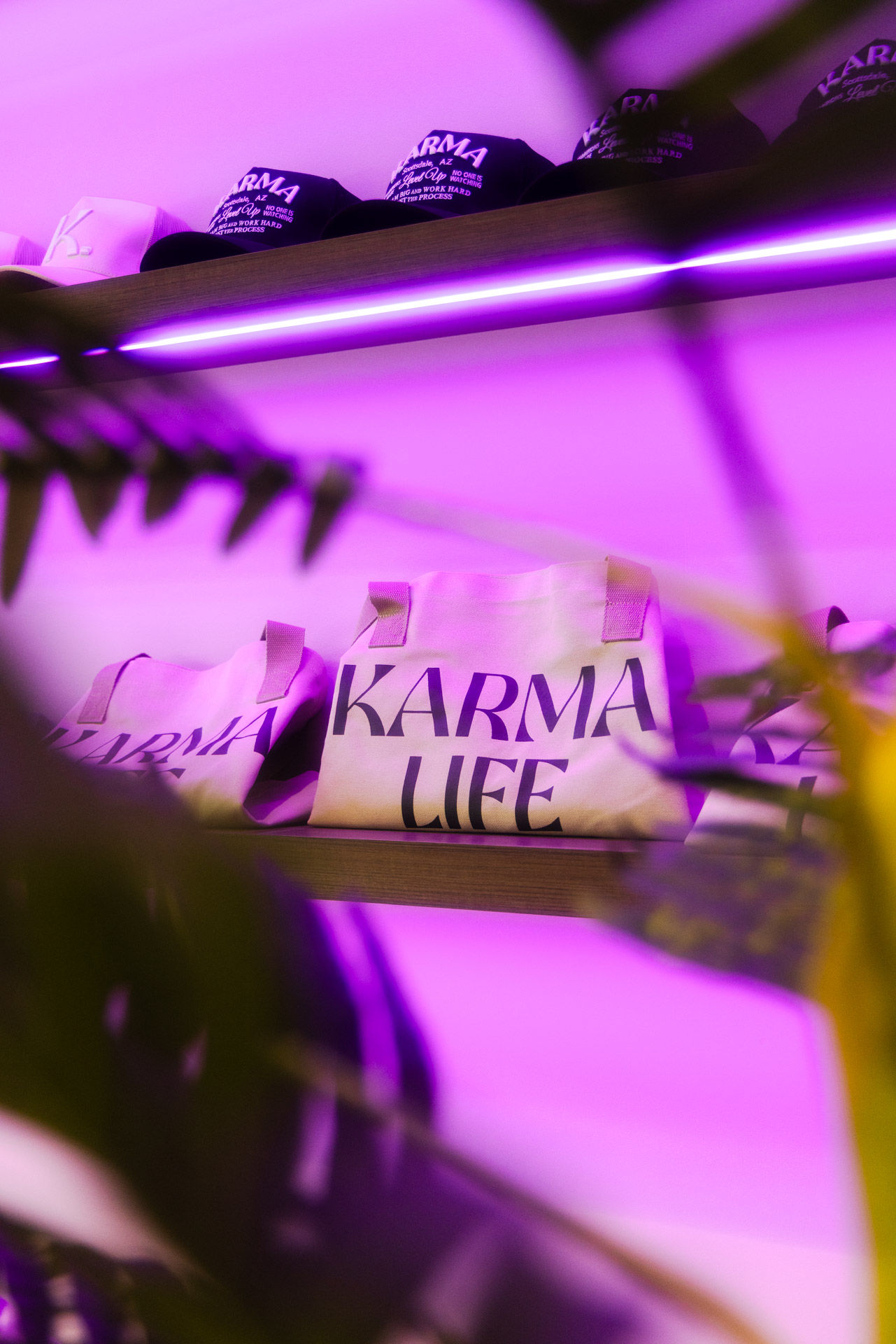 Canvas tote bag reading 'KARMA LIFE' on a wooden retail shelf with caps above, blurred plants in the foreground and vibrant purple neon lighting
