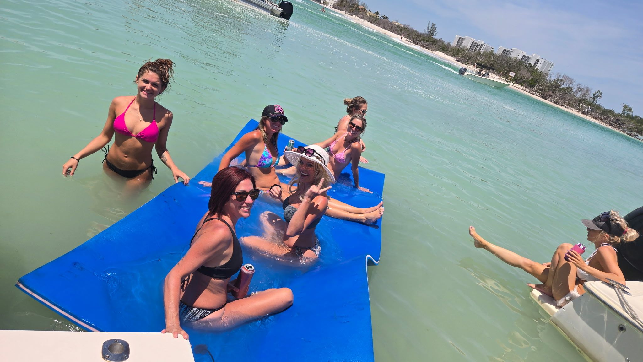 Group of women in bikinis relaxing and laughing on a large blue floating mat beside a boat in shallow turquoise water near a sandy shoreline.