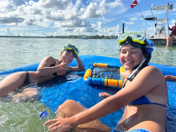 Two smiling girls in snorkel masks relaxing on a blue inflatable float in shallow clear coastal water on a sunny day, with a small boat and dive flag in the background.
