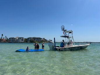 Group of people on a blue floating mat next to a center-console fishing boat in clear shallow turquoise water with waterfront homes and a bright blue sky.