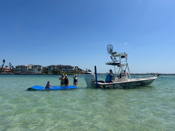 Group of people on a blue floating mat next to a center-console fishing boat in clear shallow turquoise water with waterfront homes and a bright blue sky.