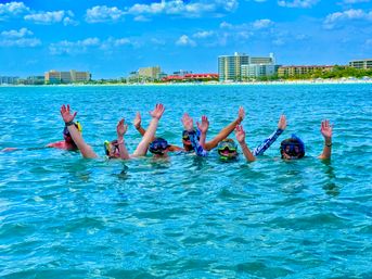 Group of snorkelers in masks and snorkels waving while floating in turquoise water with a sandy beach and coastal resort hotels on the shoreline under a blue sky