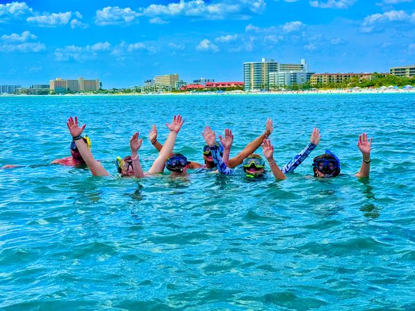 Group of snorkelers in masks and snorkels waving while floating in turquoise water with a sandy beach and coastal resort hotels on the shoreline under a blue sky