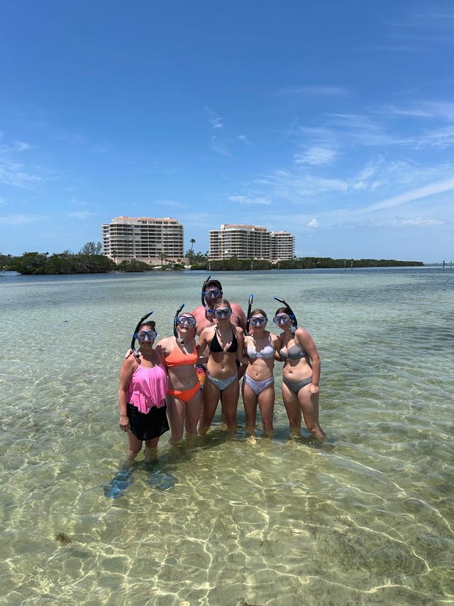 Six snorkelers posing in crystal-clear shallow coastal water near an inlet, with beachfront condominium buildings and a bright blue sky overhead