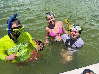 Three snorkelers in green coastal water at Siesta Key Florida, smiling and holding a large conch shell — bright rash guard, pink bikini, and snorkel masks visible.