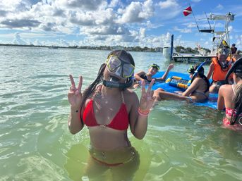 Person in red bikini wearing a snorkel mask flashes peace signs while waist‑deep in clear tropical bay water, friends on a blue float and a boat with a dive flag nearby under a sunny sky.